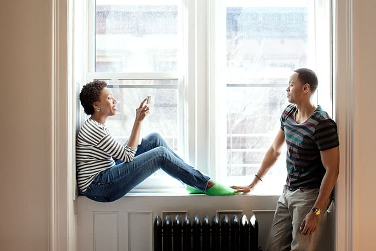 Frau sitzt auf der Fensterbank über einer Heizung und unterhält sich mit Mann