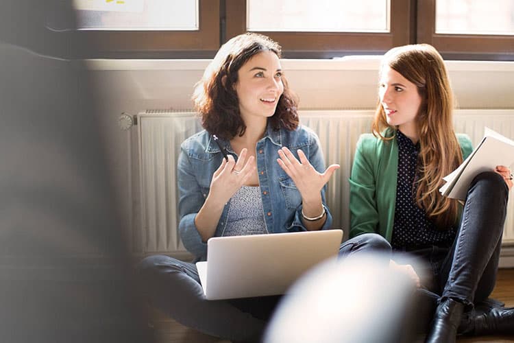 Zwei Frauen sitzen mit Laptop und Unterlagen auf dem Boden vor einer Heizung