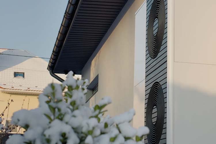 Wärmepumpe an einer Hauswand, davor ein eingeschneiter Busch