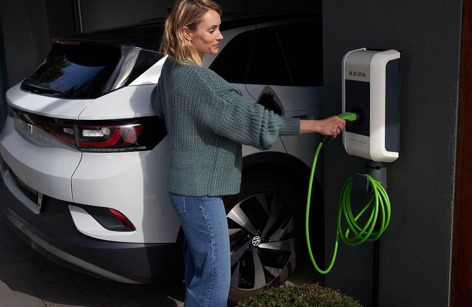 Frau bedient Wallbox an Hauswand neben einem E-Auto.