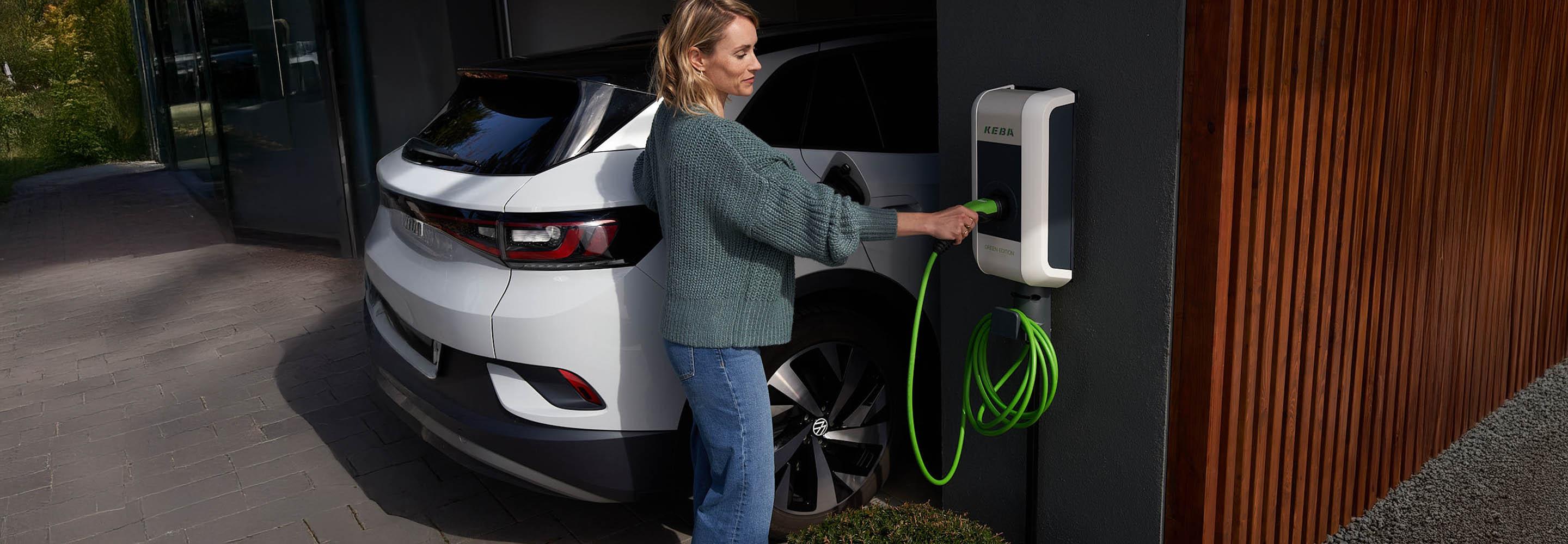 Frau bedient Wallbox an Hauswand neben einem E-Auto.
