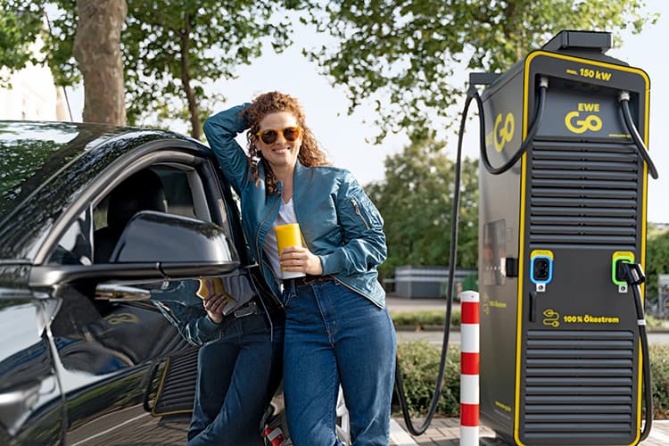 Eine Frau lehnt an ihrem E-Auto während es an einer EWE Go Ladestation lädt. 