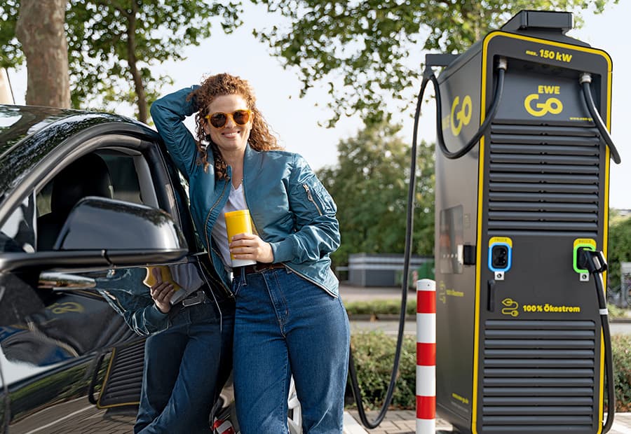 Eine Frau lehnt sich mit einem Kaffeebecher an ihr E-Auto, das an einer EWE GO Ladesäule lädt. 