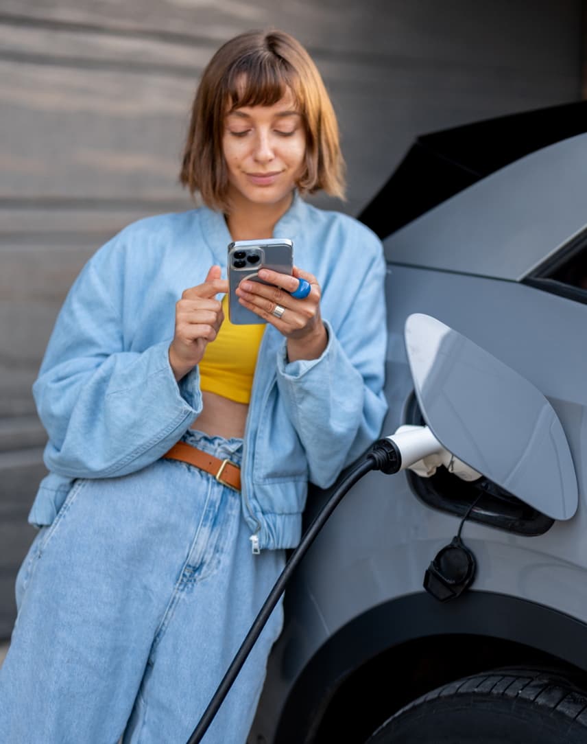 Frau bedient Handy angelehnt an einem E-Auto.