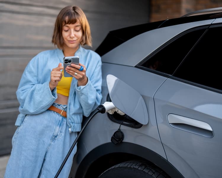 Frau bedient Handy angelehnt an einem E-Auto.
