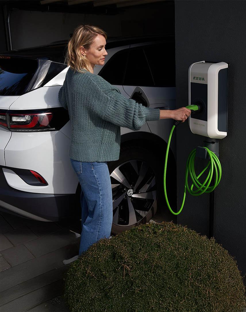 Frau bedient Wallbox an Hauswand neben einem E-Auto.