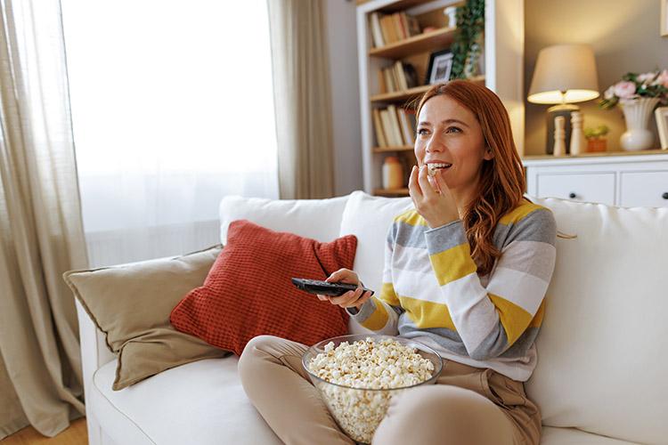Frau sitzt auf dem Sofa und isst Popcorn mit Fernbedienung in der Hand