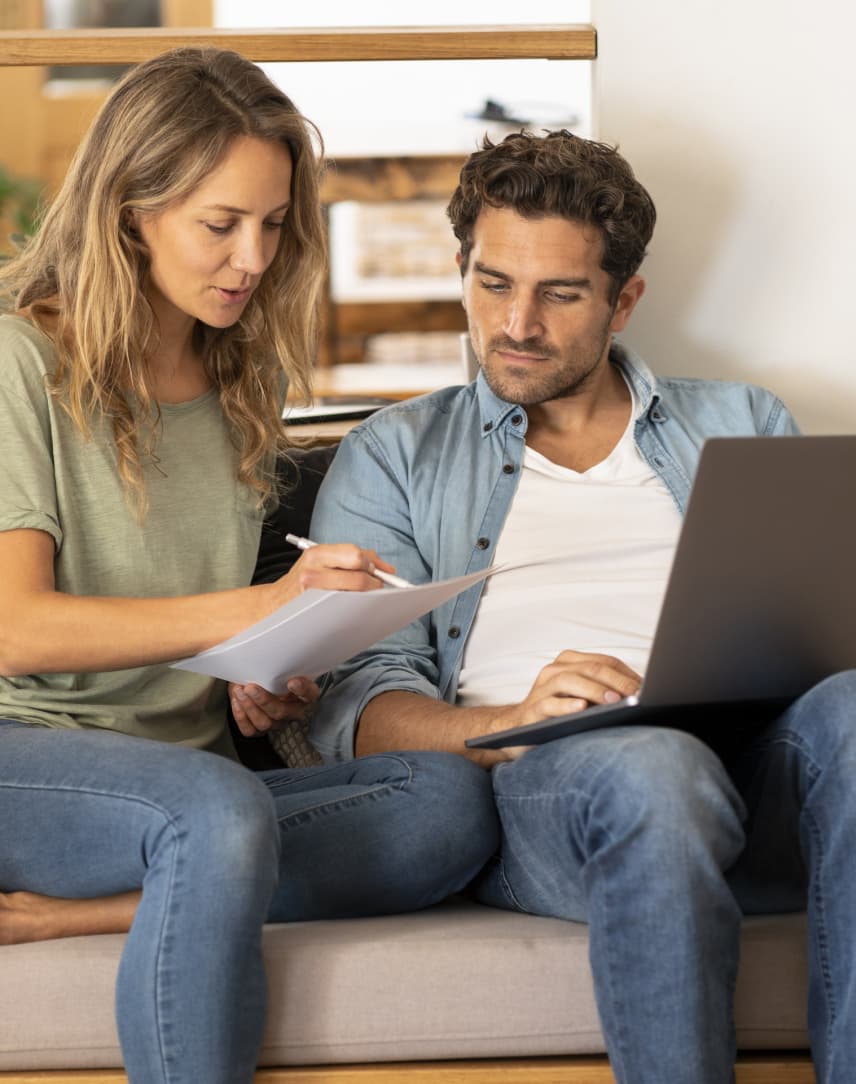 Ein Paar sitzt auf der Couch, schaut sich Dokumente an und schreibt dabei auf einem Laptop