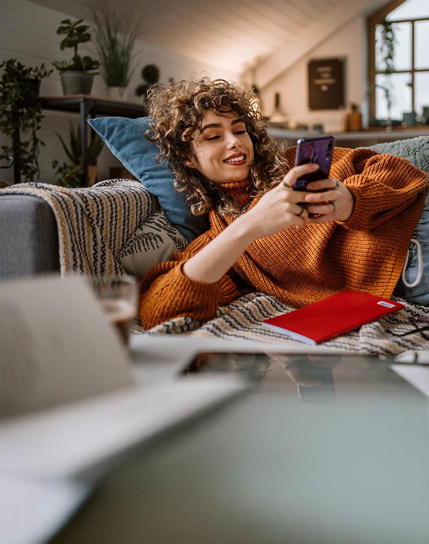 Eine Frau liegt entspannt auf dem Sofa und schaut auf ihr Smartphone