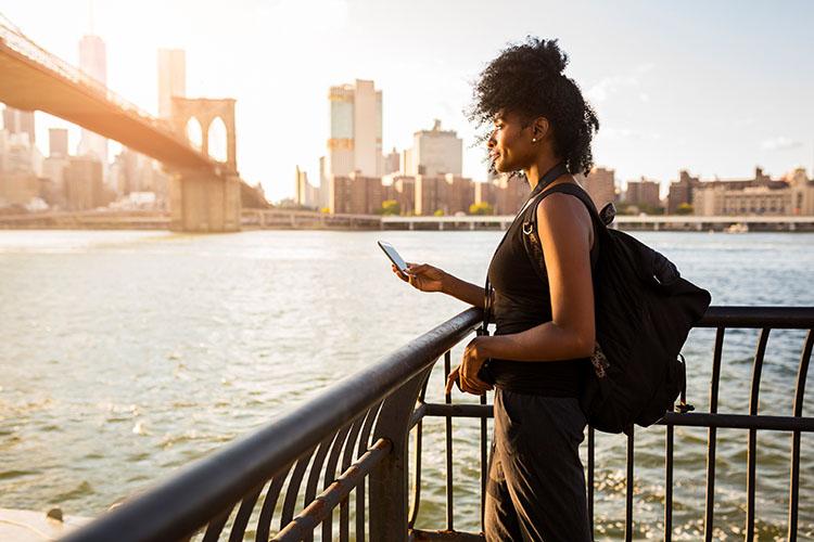 Frau mit Handy in der Hand schaut am Wasser auf New York City.