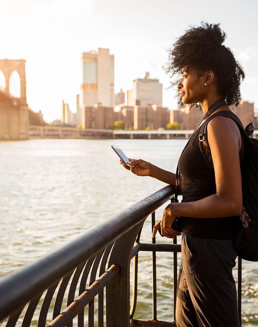 Frau mit Handy in der Hand schaut am Wasser auf New York City.