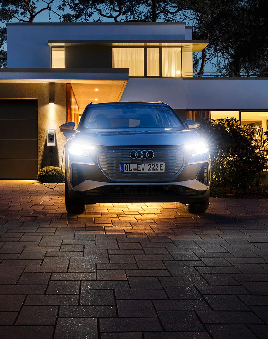 Weißes Auto vor beleuchtetem Haus an einer Wallbox angeschlossen