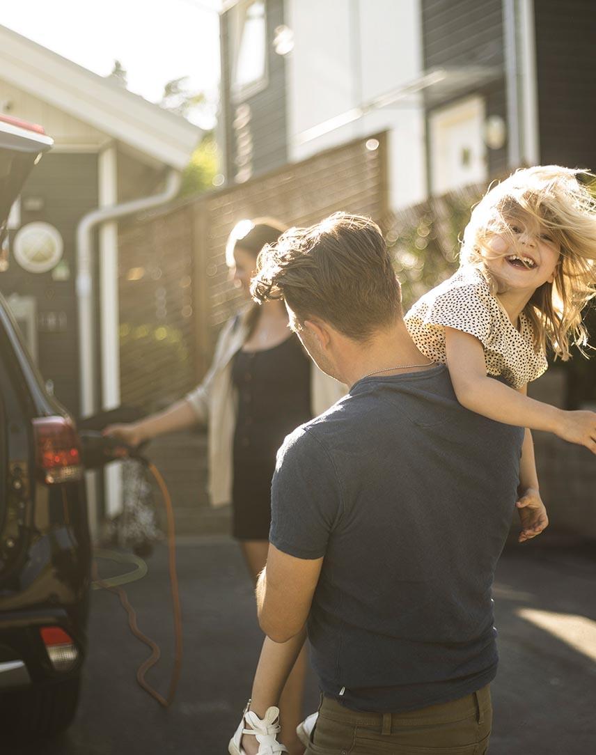 Mann trägt Tochter über der Schulter, im Hintergrund lädt eine Frau ein E-Auto