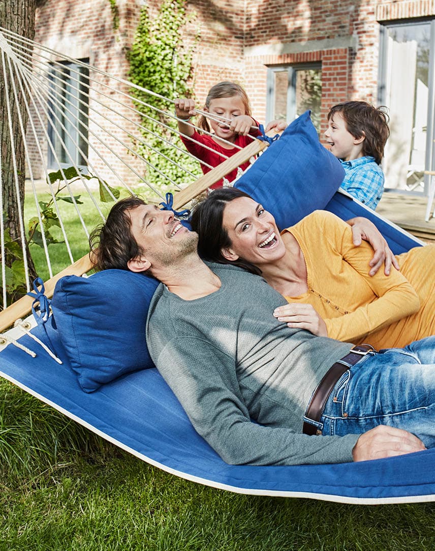 Mutter und Vater liegen lachend in einer Hängematte, ihre Kinder spielen daneben