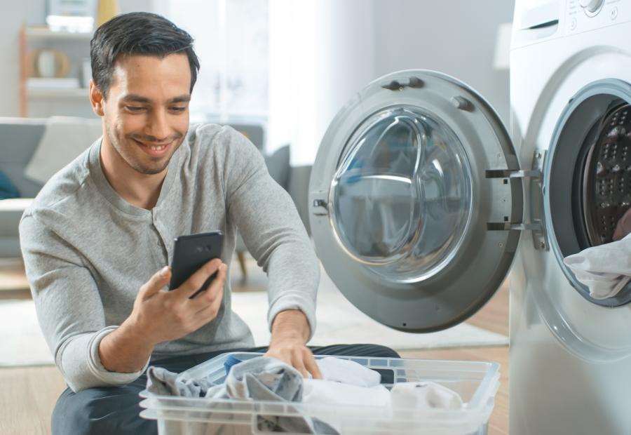 Mann kniet vor einer Waschmaschine und schaut auf sein Smartphone