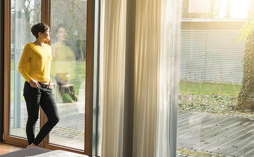 Frau steht am Fenster und schaut in ihren sonnendurchfluteten Garten