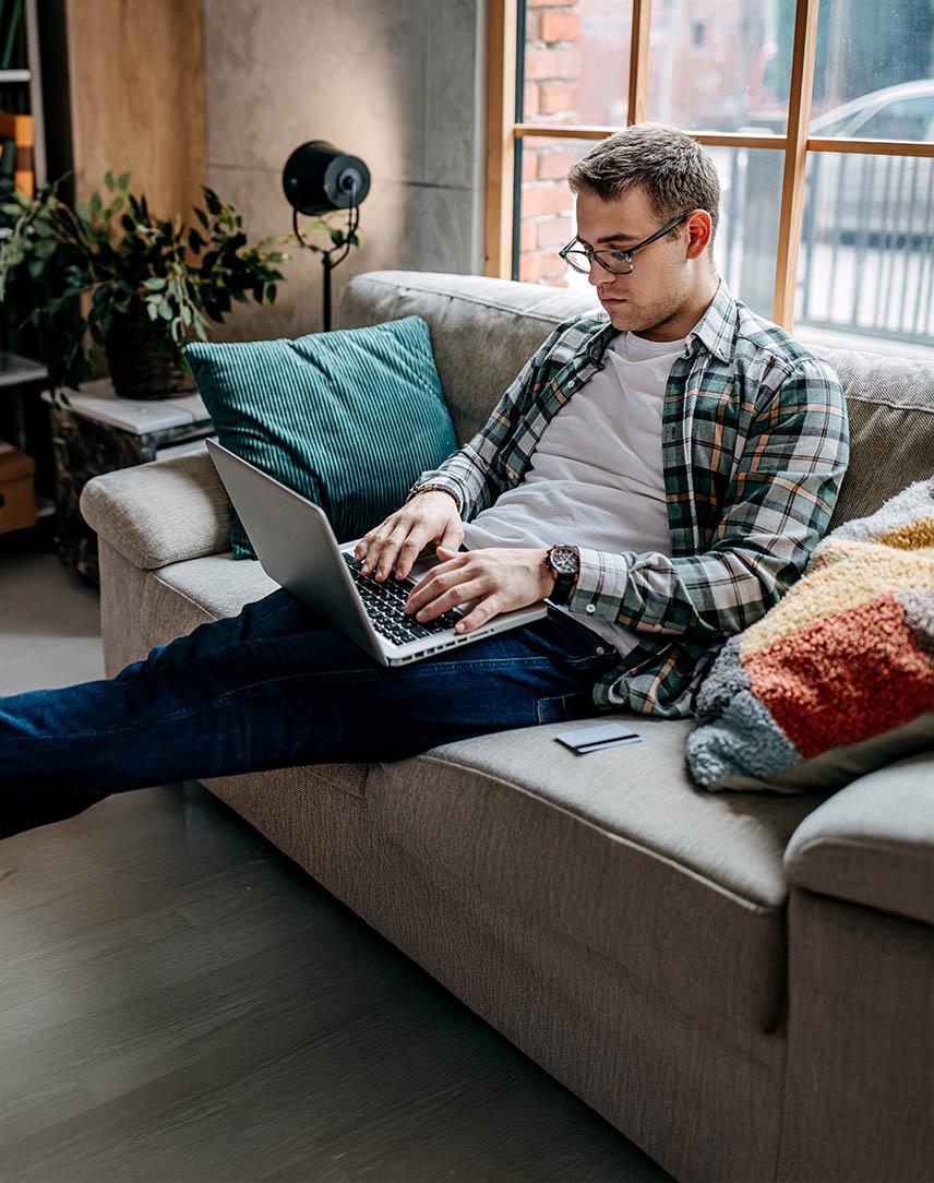 Junger Mann sitzt auf der Couch beim Fernstudium an seinem Laptop