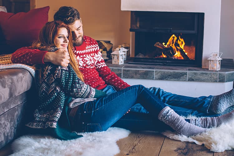 Pärchen sitzt Arm in Arm auf einem Fell vor dem laufenden Kamin