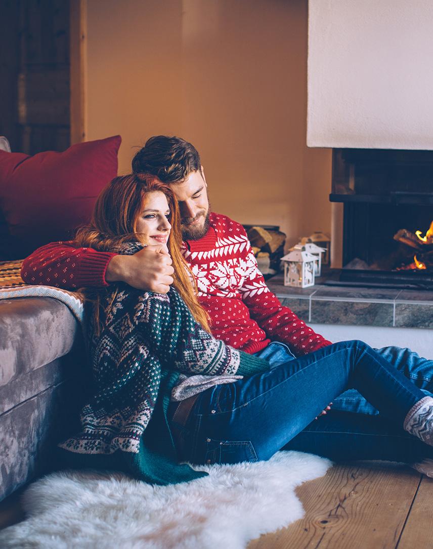 Pärchen sitzt Arm in Arm auf einem Fell vor dem laufenden Kamin