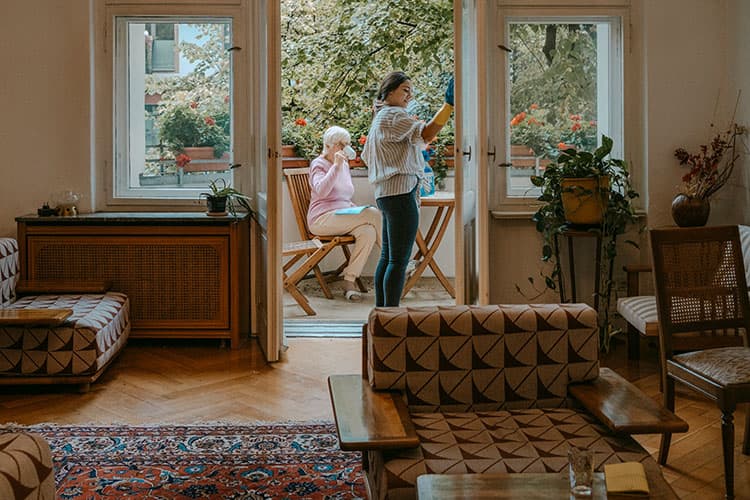 Ältere Dame trinkt auf dem Balkon Kaffee während eine andere Frau Fenster putzt