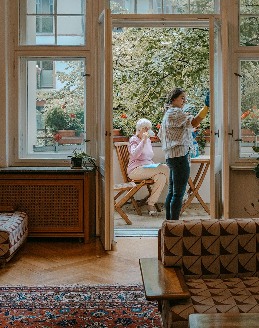 Ältere Dame trinkt auf dem Balkon Kaffee während eine andere Frau Fenster putzt