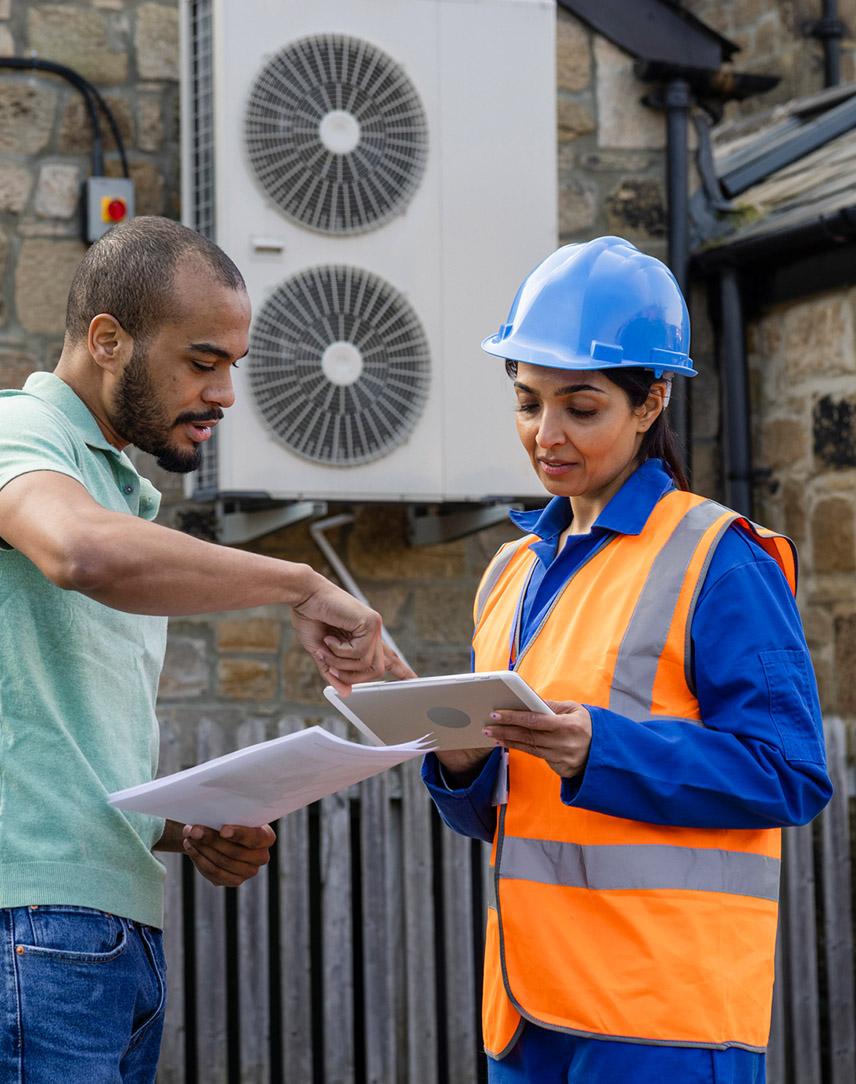 Frau mit Warnweste und Bauhelm prüft unterlagen zusammen mit Mann vor Wärmepumpe