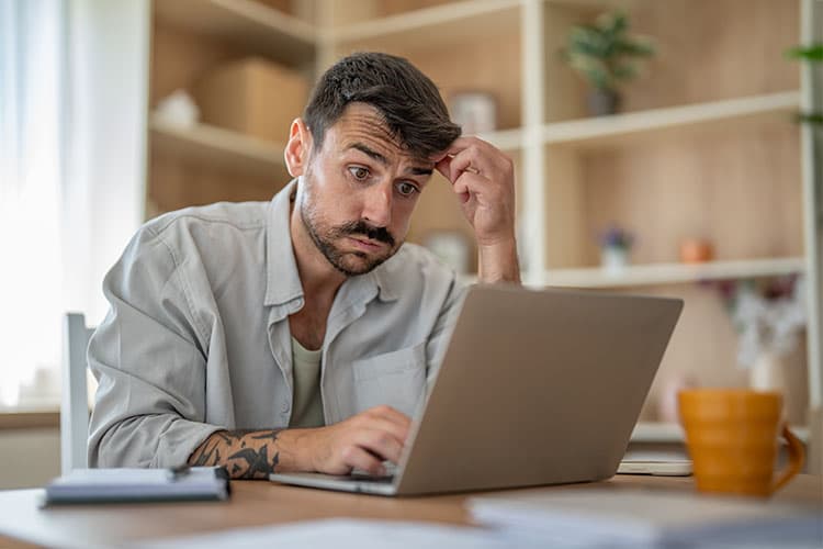 Mann mit nachdenklichem Gesichtsausdruck sitzt vor einem Laptop und berührt seine Stirn