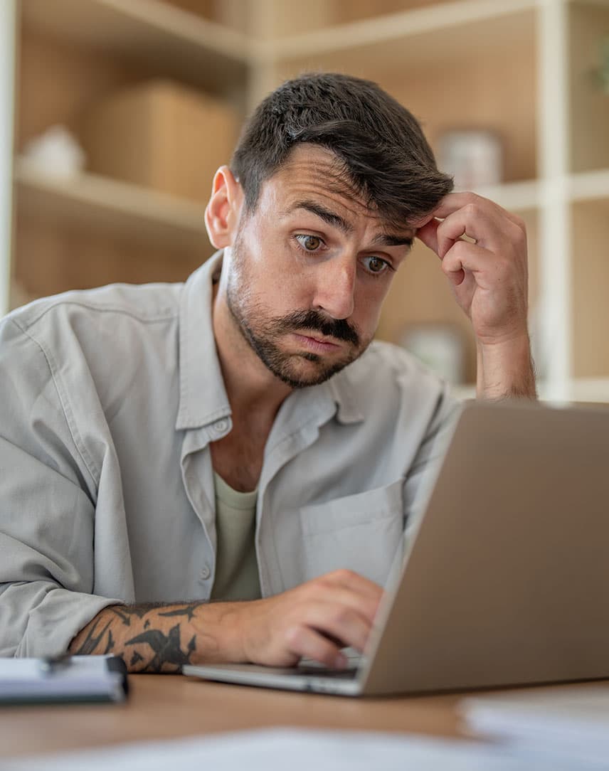 Mann mit nachdenklichem Gesichtsausdruck sitzt vor einem Laptop und berührt seine Stirn