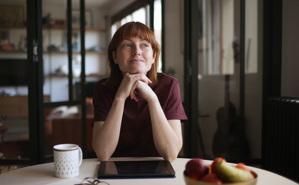 Eine rothaarige Frau sitzt an einem Tisch und überlegt.