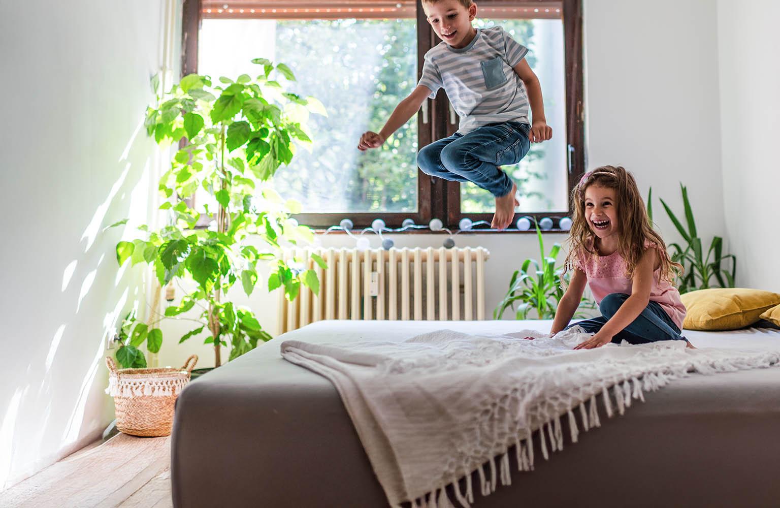 Ein kleiner Junge springt vergnügt auf einem Bett, ein kleines Mädchen schaut ihm lachend zu