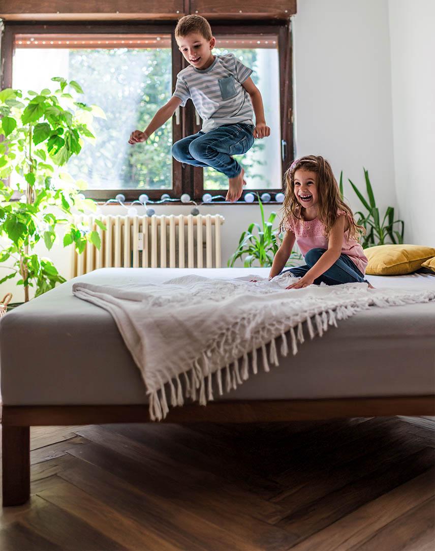 Ein kleiner Junge springt vergnügt auf einem Bett, ein kleines Mädchen schaut ihm lachend zu