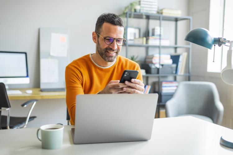 Mann sitzt vor seinem Laptop und schaut auf sein Smartphone