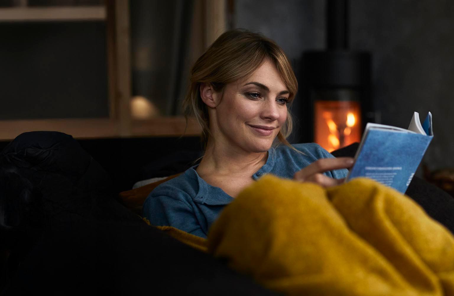 Eine Frau sitzt entspannt auf dem Sofa und liest, im Hintergrund brennt der Kamin