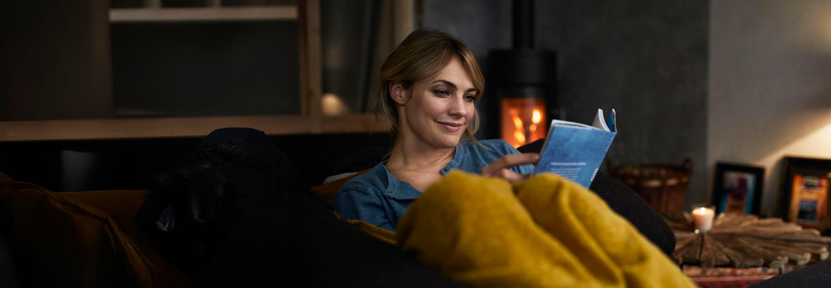 Eine Frau sitzt entspannt auf dem Sofa und liest, im Hintergrund brennt der Kamin