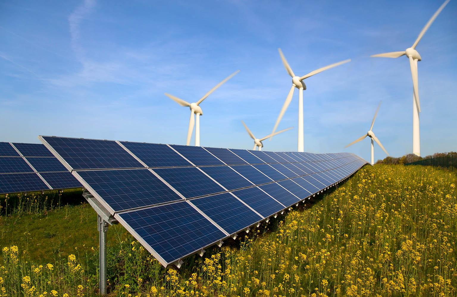 Im Vordergrund sieht man Solarmodule, im Hintergrund Windräder vor blauem Himmel