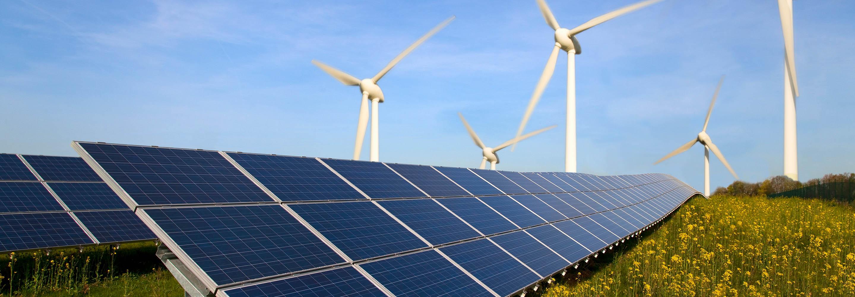 Im Vordergrund sieht man Solarmodule, im Hintergrund Windräder vor blauem Himmel