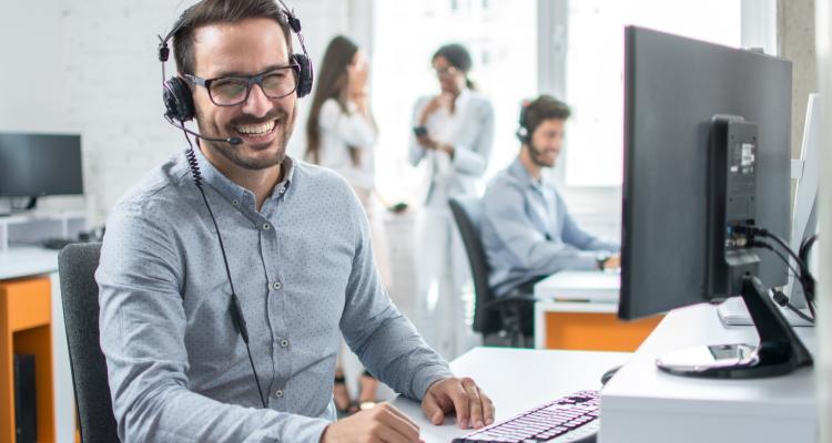 Mann mit Headset sitzt vor einem Laptop und lächelt freundlich in die Kamera