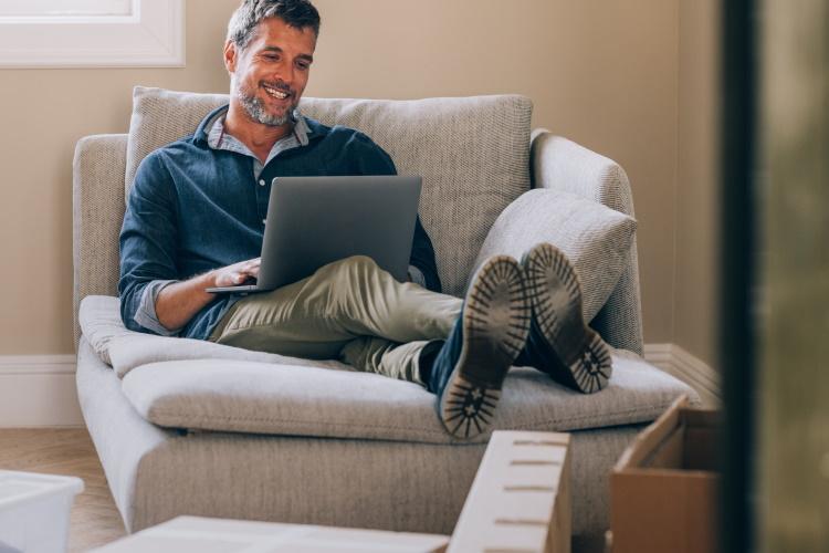 Mann sitzt inmitten von Umzugskartons auf der Couch mit einem Laptop auf den Beinen