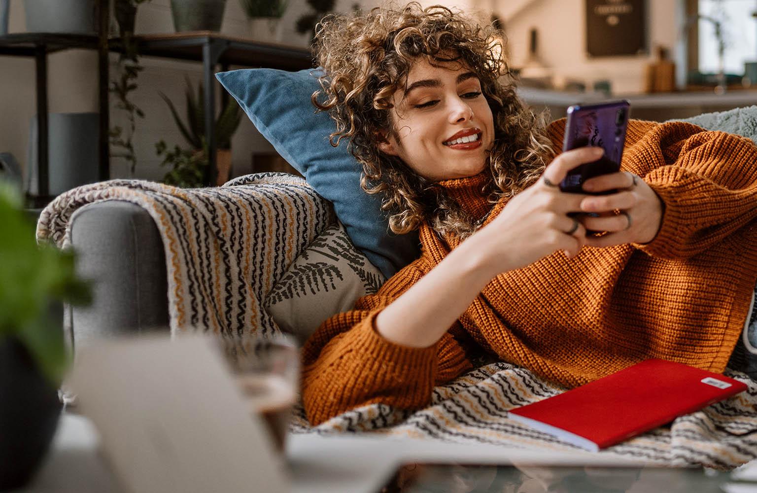 Eine Frau liegt entspannt auf dem Sofa und schaut auf ihr Smartphone