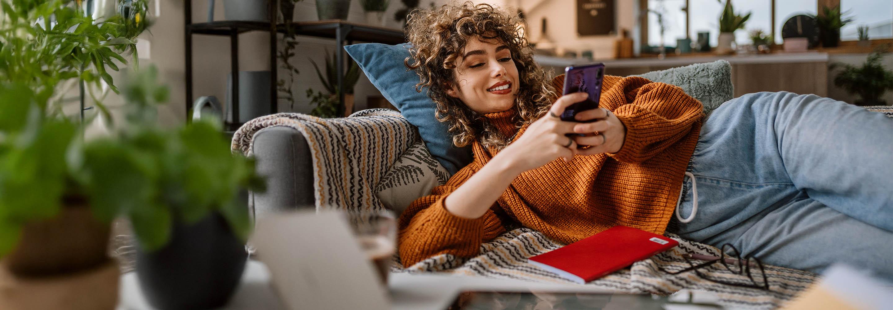 Eine Frau liegt entspannt auf dem Sofa und schaut auf ihr Smartphone