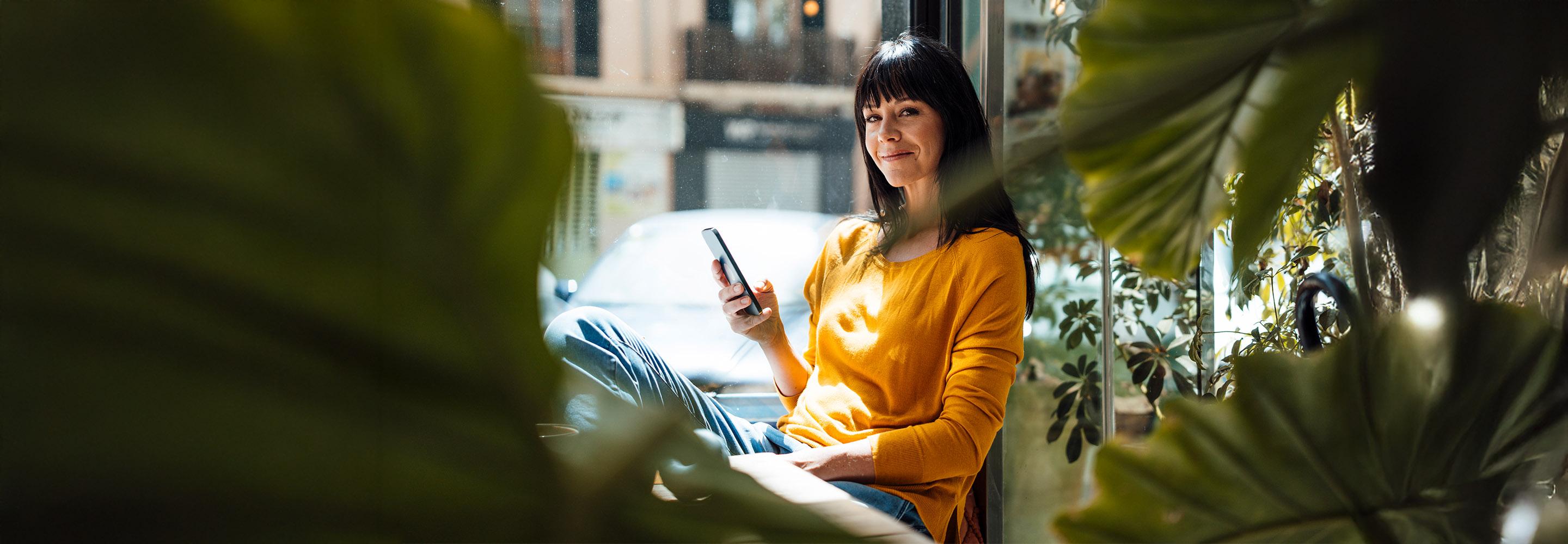 Lächelnde Frau sitzt auf Fensterbank mit Handy in der Hand