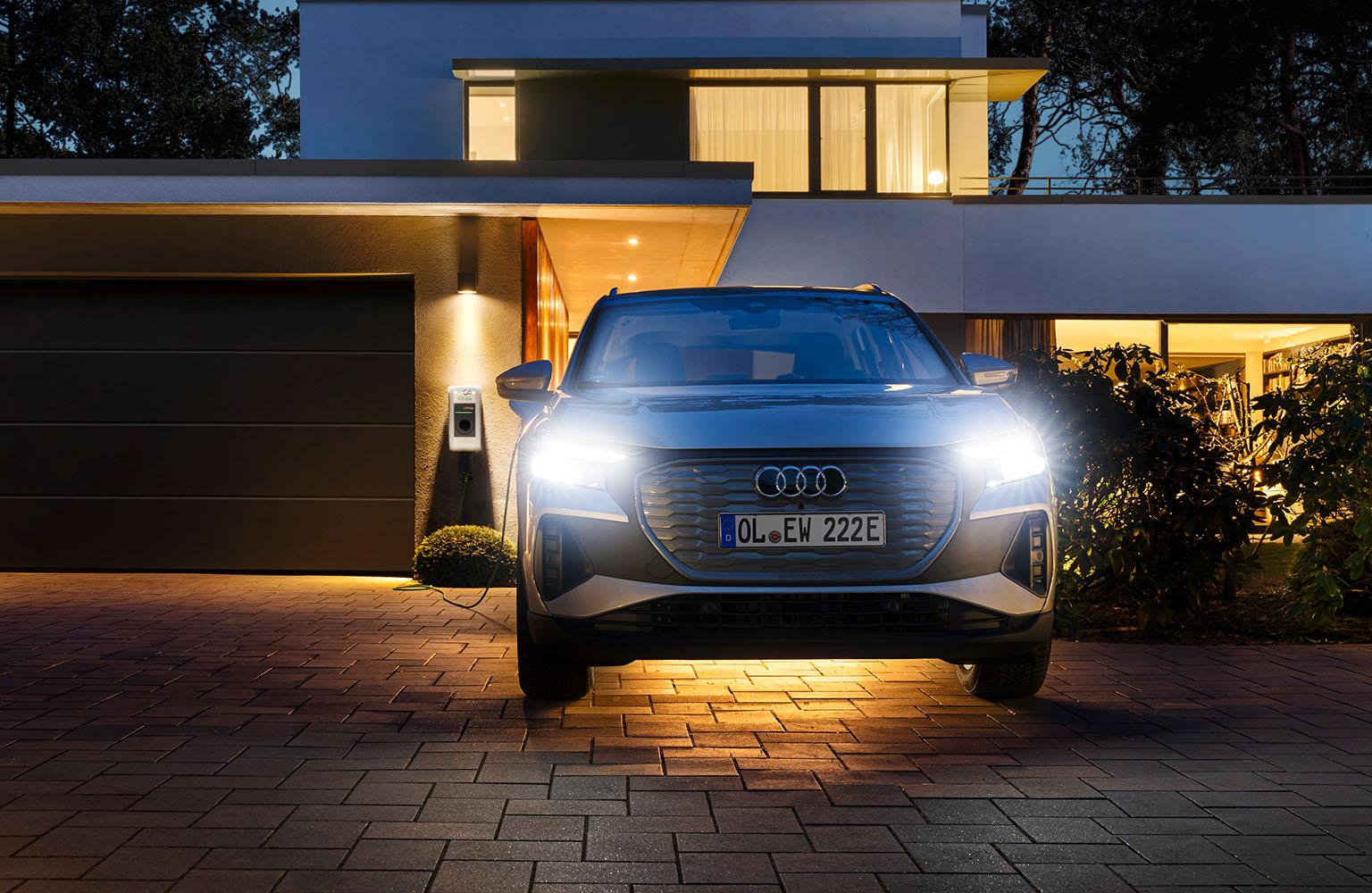 Weißes Auto vor beleuchtetem Haus an einer Wallbox angeschlossen