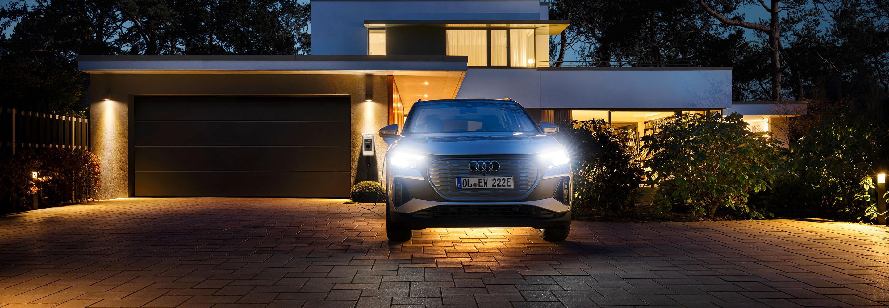 Weißes Auto vor beleuchtetem Haus an einer Wallbox angeschlossen