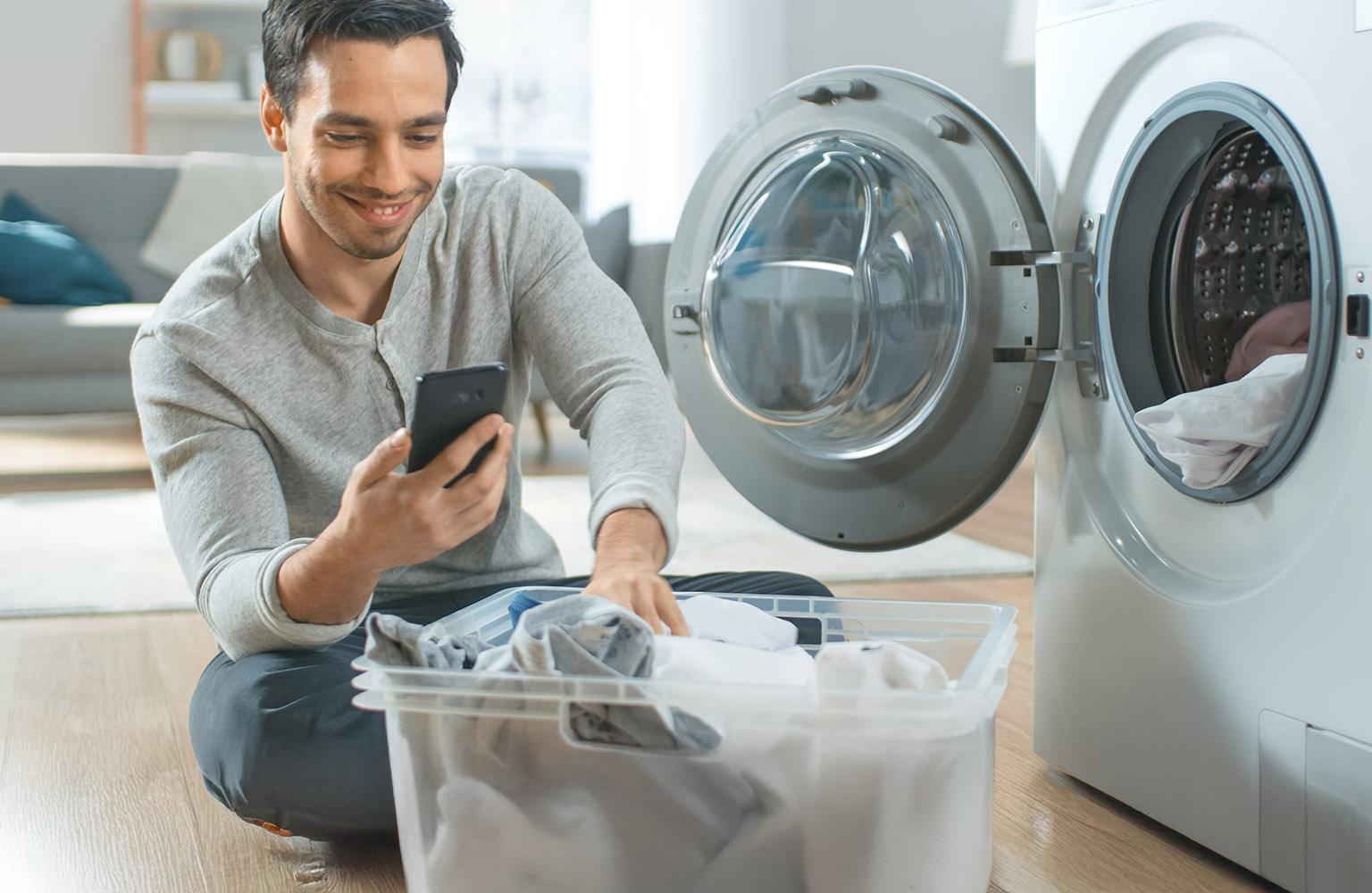 Mann mit Wäschekorb vor geöffneter Waschmaschine schaut auf sein Smartphone
