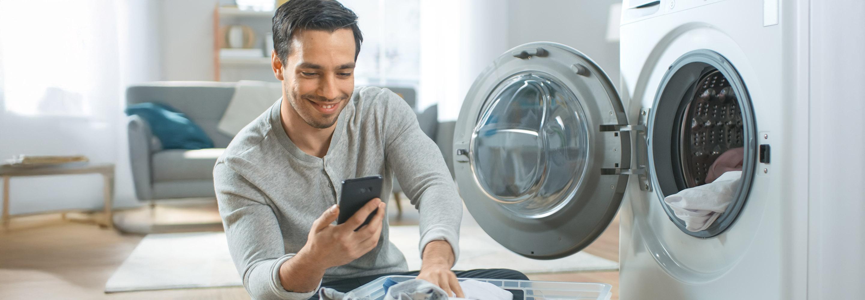 Mann mit Wäschekorb vor geöffneter Waschmaschine schaut auf sein Smartphone