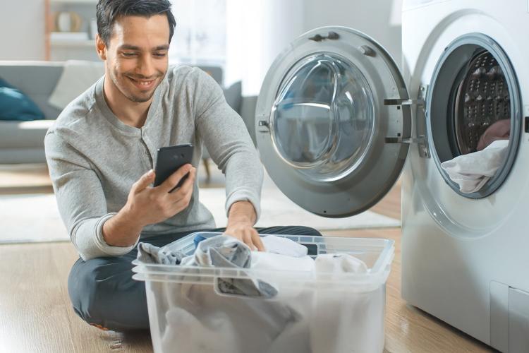 Mann mit Wäschekorb vor geöffneter Waschmaschine schaut auf sein Smartphone