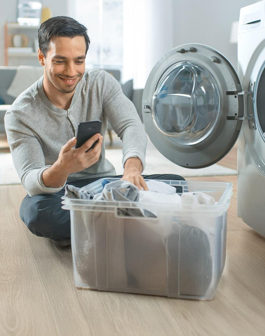 Mann mit Wäschekorb vor geöffneter Waschmaschine schaut auf sein Smartphone