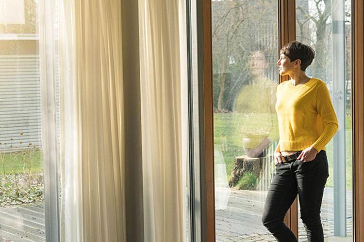 Frau steht am Fenster und schaut in ihren sonnendurchfluteten Garten