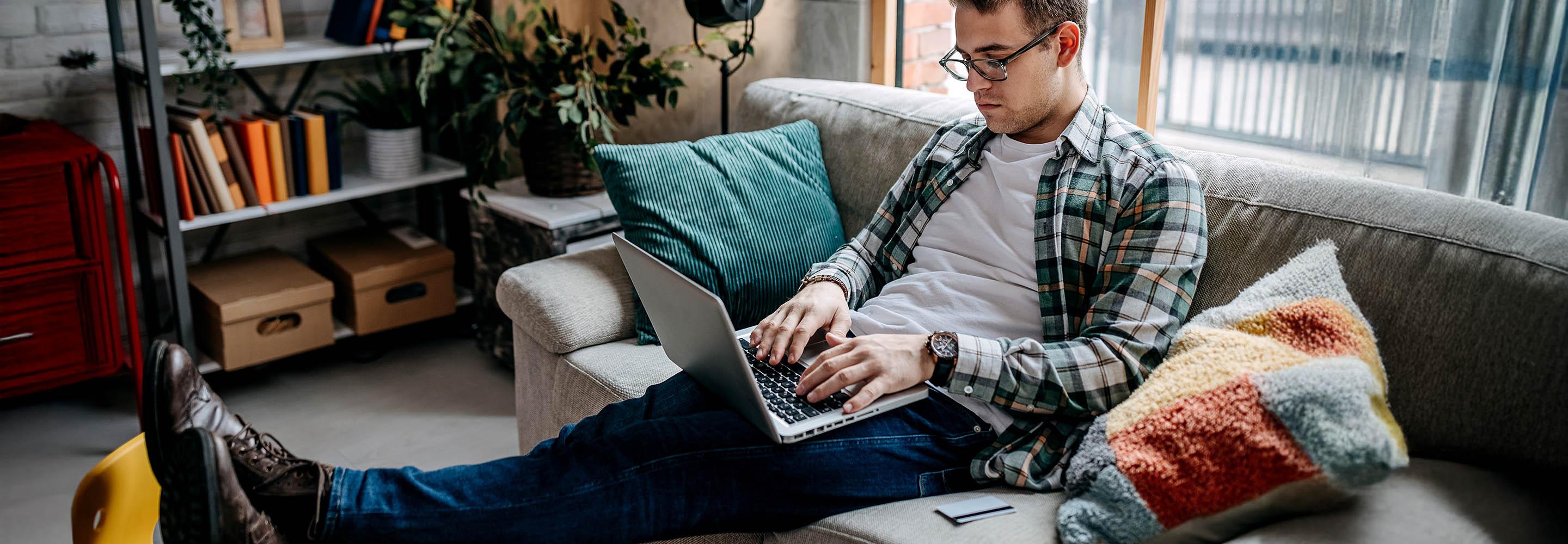 Junger Mann sitzt auf der Couch beim Fernstudium an seinem Laptop