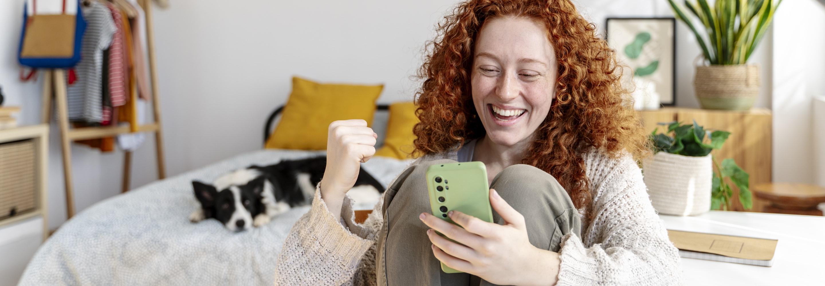 Rothaarige Frau am Schreibtisch schaut auf ihr Smartphone und freut sich